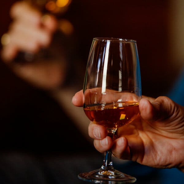 A hand holds up a glass partially filled with an amber liquid. 