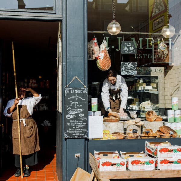 A woman places a bread in a shop window, while another employee in a white shirt and a brown apron stands in the doorway. 