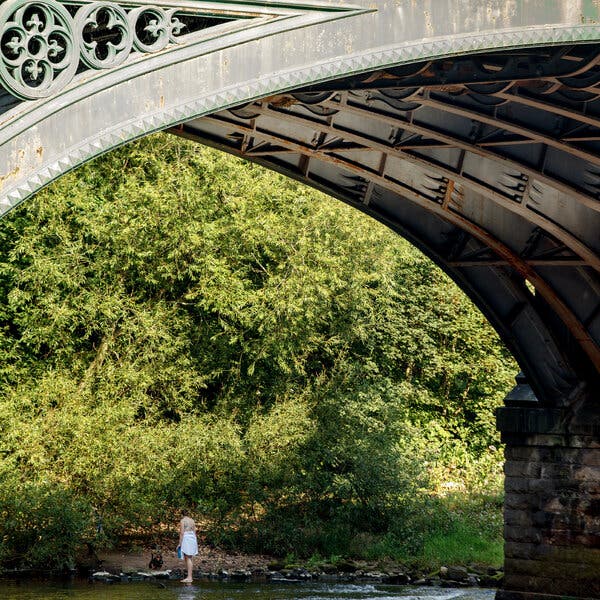 A woman stands in shallow water beneath the curve of a stone bridge above. 