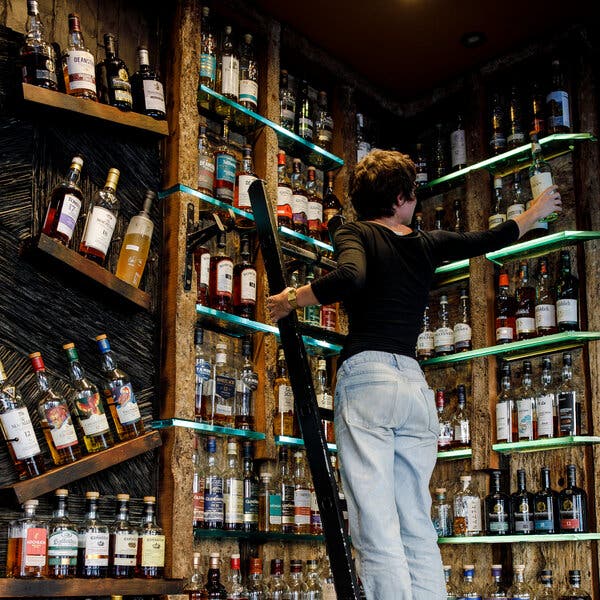 A woman with her back to the camera stands on a ladder to take a bottle of alcohol off a shelf. 