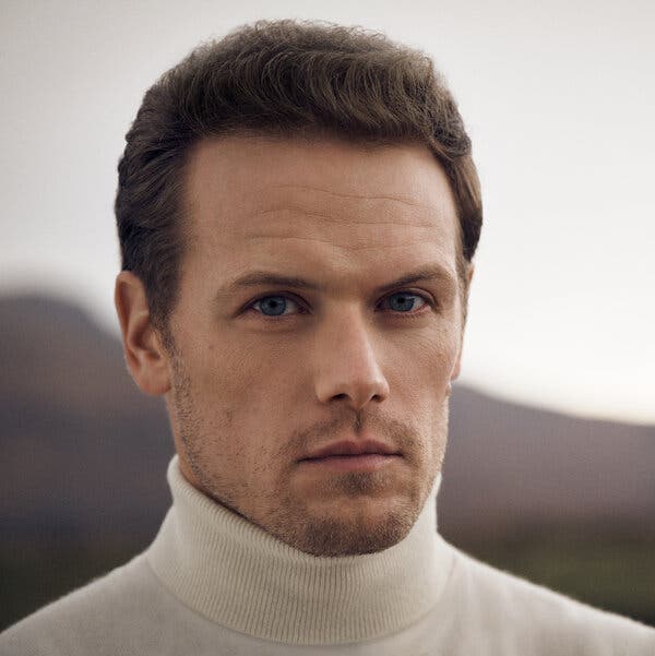 Sam Heughan looks straight into the camera. He has dark brown hair, a stubble beard and is wearing a white turtleneck. 