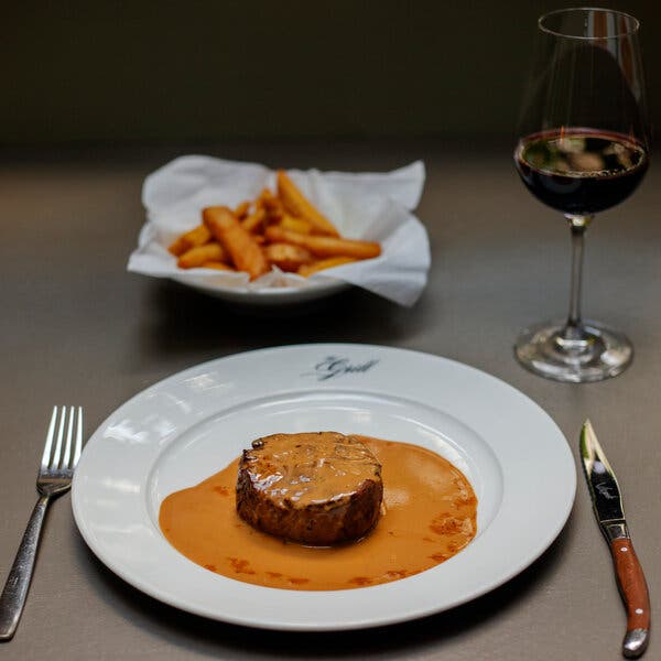 A filet of meat sits on a white plate covered in an orange sauce. A dish of fries is behind it on the table. 