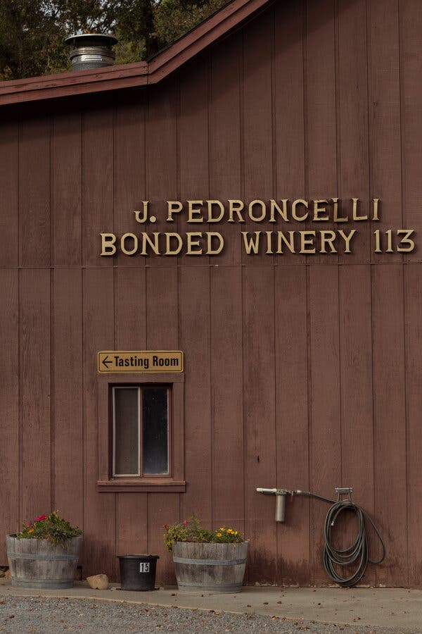 A brown wooden building with a small window and two planters and a hose on the wall has letters reading, "J. Pedroncelli, Bonded Winery 113" and a sign that says "Tasting Room" with an arrow pointing to the left.