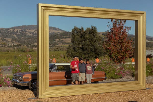 Three men stand next to a sleek, brown classic car in the center of a large, gold picture frame sculpture. There are green fields, rolling hills, trees and flowers behind them along a two-lane highway.