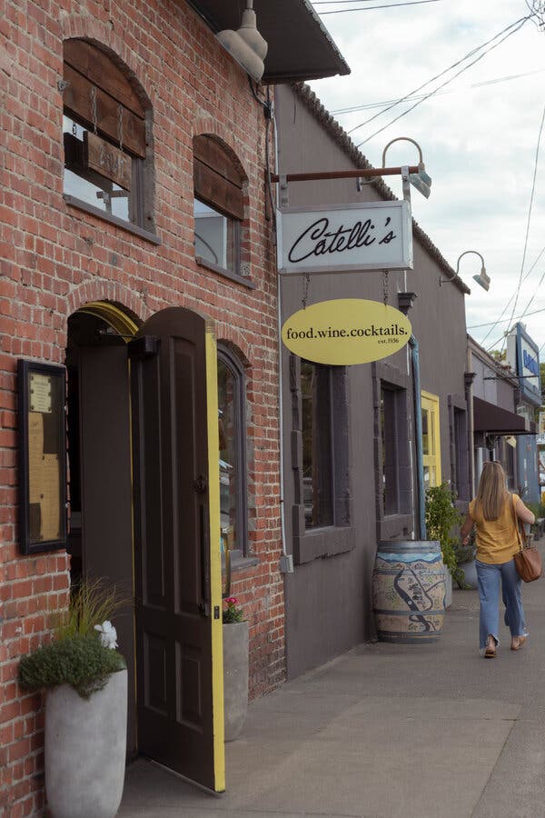 A red brick storefront with arched windows and doors and two cylindrical concrete planters around the entrance. Signs hanging above the sidewalk read “Catelli's” and “Food, Wine, Cocktails, Est. 1936.”