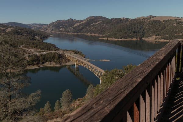 A view of a long, smooth lake surrounded by olive green hills covered with trees and bushes. A steel bridge crosses the lake at a narrow point, and the sky is cloudless.