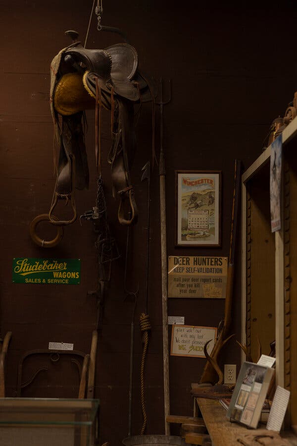 A saddle hangs from the ceiling in a room that has signs advertising wagons and addressed at deer hunters, as well as a shelf with photos and other curios on it.