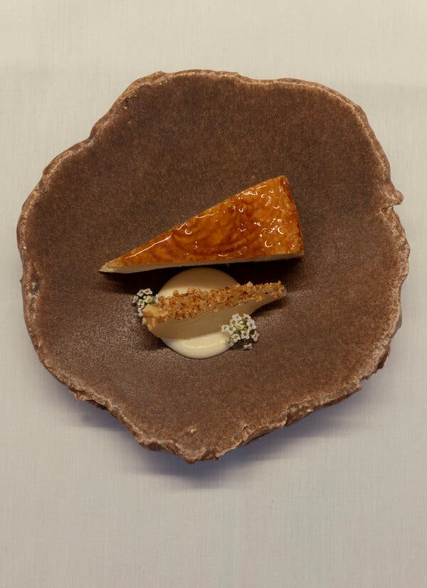 A shiny wedge of cake sits next to a creamy dollop decorated with tiny, white flowers in a rough, brown bowl.