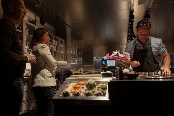 A man wearing a short-sleeved blue-gray shirt and a dark striped apron stands behind a counter that has brown bowls, silver trays, and a bouquet of pink and white cosmos on it. To the left, two people stand watching him work, one holding a glass of white wine.